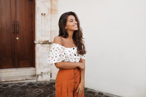 Tanned woman in stylish orange shorts with high waist and light blouse posing against background of house with antique wooden doors