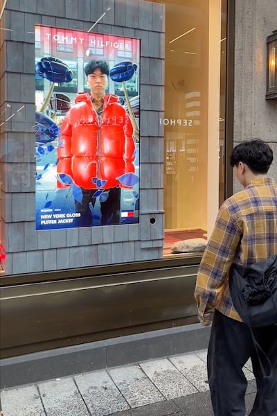 The image depicts a person standing with their back to the camera, wearing a yellow and black plaid shirt and dark pants. They carry a black backpack. The focal point of the image is a Tommy Hilfiger advertisement displayed on the exterior wall of a store. The advertisement features an individual wearing a bright red puffer jacket, with the face obscured for privacy. The text on the ad reads: “NEW YORK GLOSS PUFFER JACKET.” Reflections of nearby buildings are visible on the glass covering of the ad.