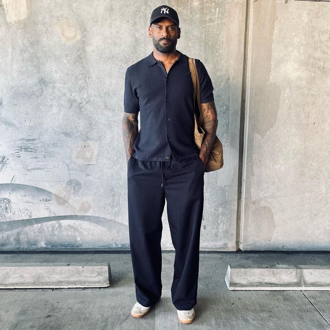 man in dark-colored short-sleeve shirt and drawstring pants. he's standing in front of a concrete wall