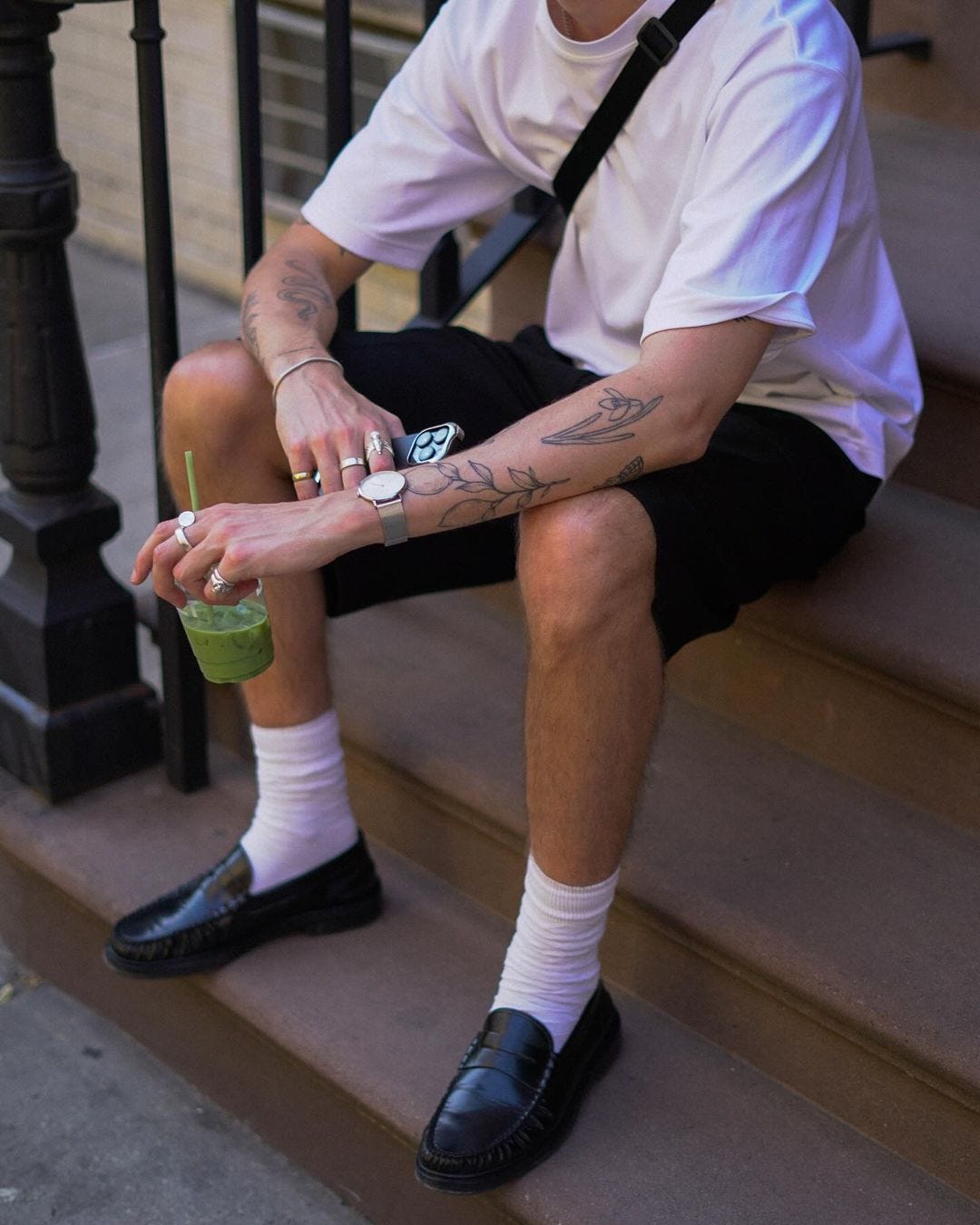 image of a man from the neck down. he's wearing a white t-shirt, black fanny pack slung over his shoulder, black shorts and white socks with black penny loafers. he's holding a green drink
