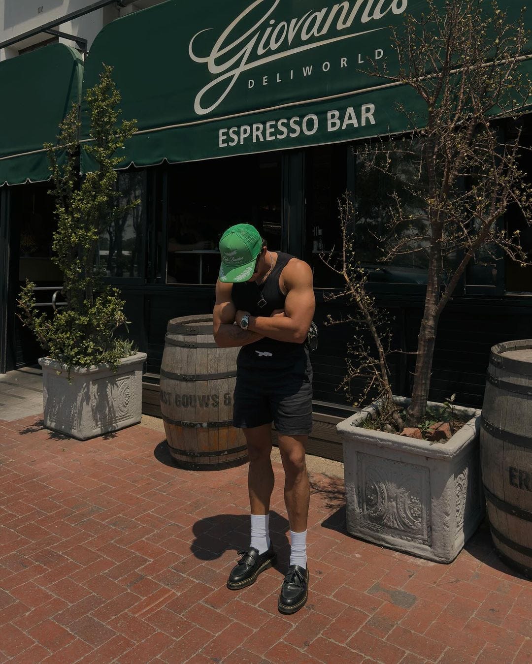 man outside in front of a bar, looking at his feet. he's wearing a green baseball cap, blank tank top and shorts, with tall white socks and loafers