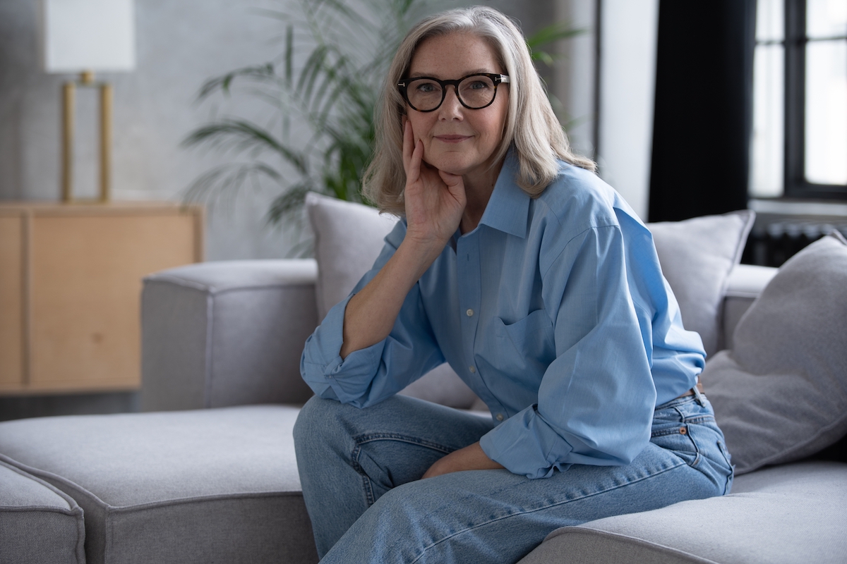 Mature woman with gray hair wearing jeans a light blue button-down shirt sits on her gray couch at home