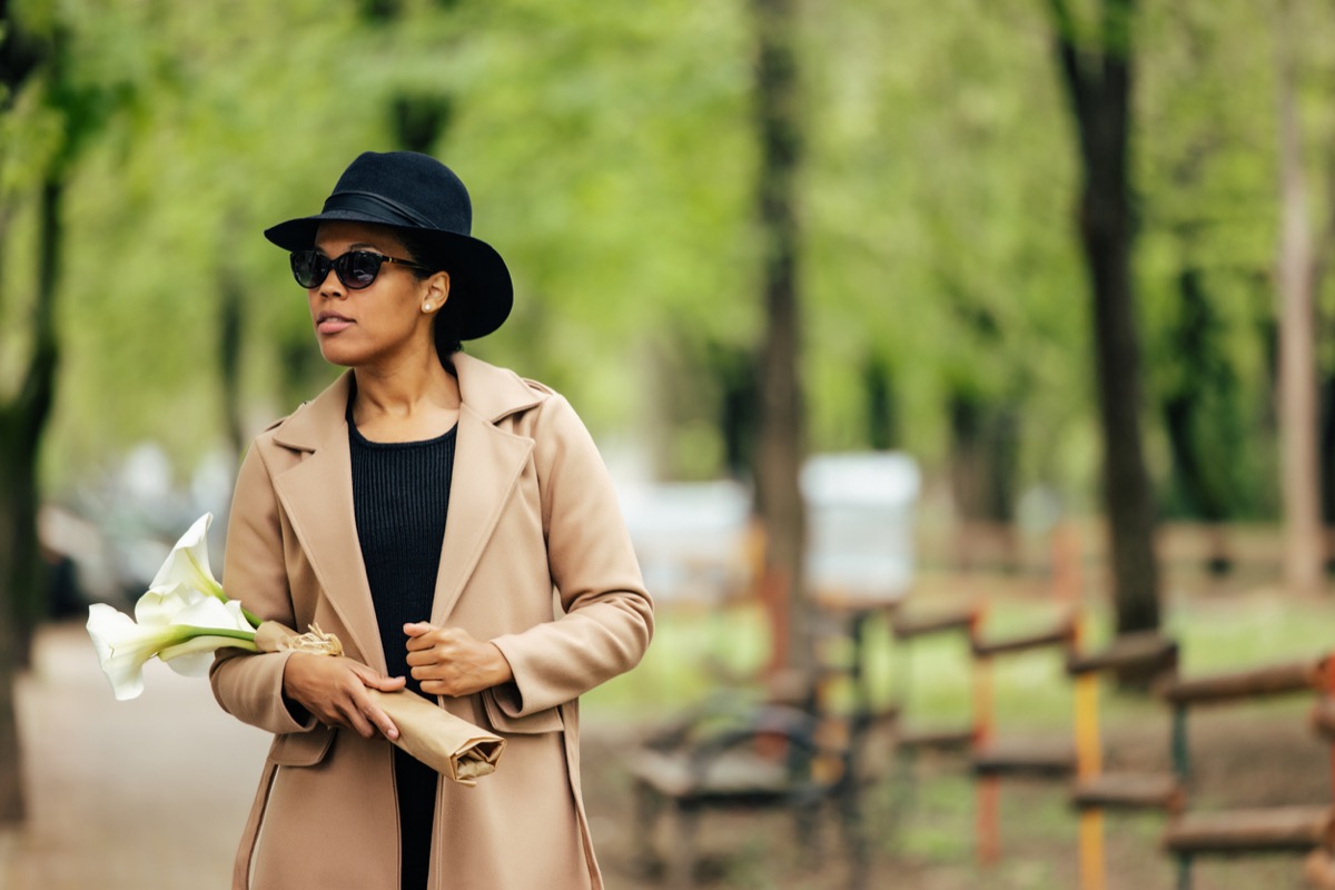 fashionable woman wearing camel coat and black fedora, carrying lilies while walking through the park