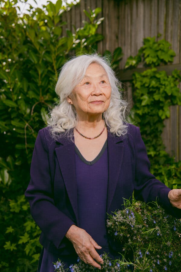 Maxine Hong Kingston poses outside her home, surrounded y greenery. She wears a deep purple knit dress and jacket. Her white hair falls over her shoulders, and she is smiling.