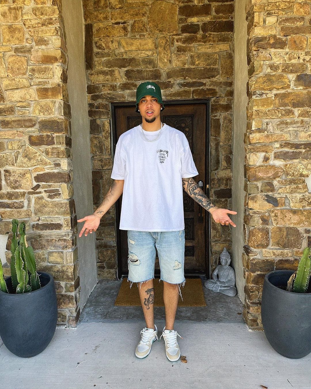 man standing in a doorway wearing a green baseball cap, white t-shirt, light ripped jean shorts, and Nike sneakers