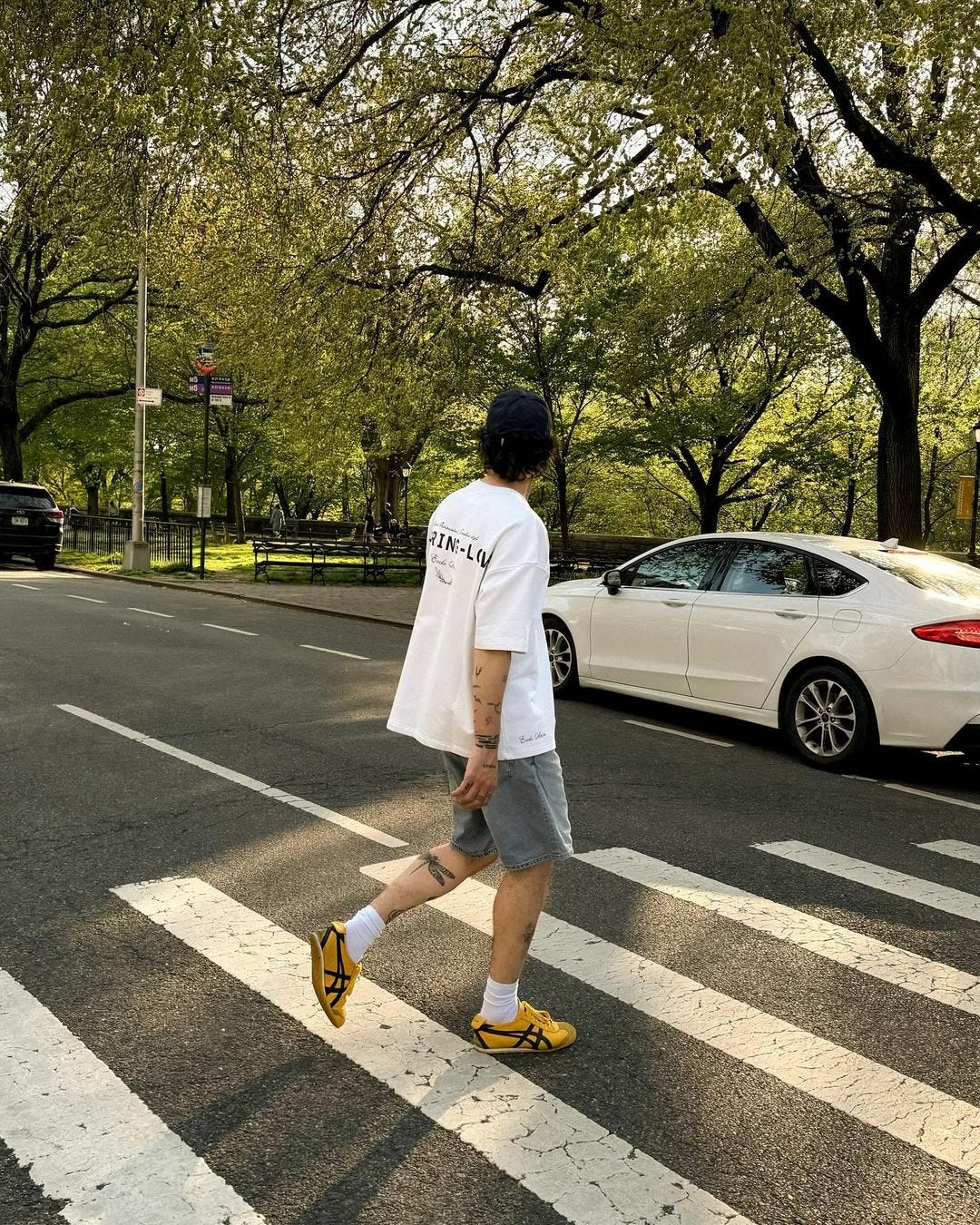 image of a man crossing the road, facing away from the camera, wearing a white t-shirt, jean shorts, tall white socks and yellow sneakers