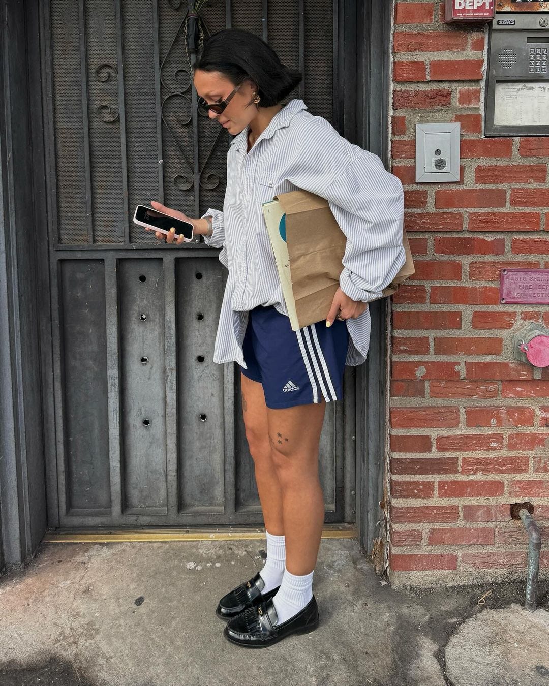 woman standing in a doorway wearing a button down shirt, blue adidas shorts, white socks, and dark loafers