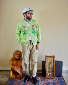 man in light green cardigan with pink embroidered detail on the chest with khaki-colored drawstring pants and chunky loafers. he's looking to the side and is also wearing a baseball hat