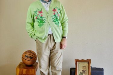 man in light green cardigan with pink embroidered detail on the chest with khaki-colored drawstring pants and chunky loafers. he's looking to the side and is also wearing a baseball hat