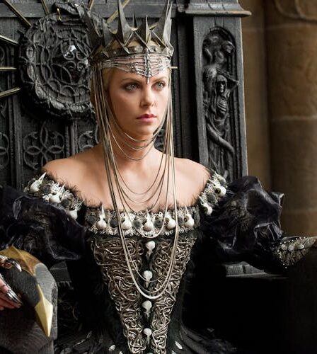 Charlize Theron playing a wicked queen in the film Snow White and the Huntsman sitting upon a throne wearing a crown.