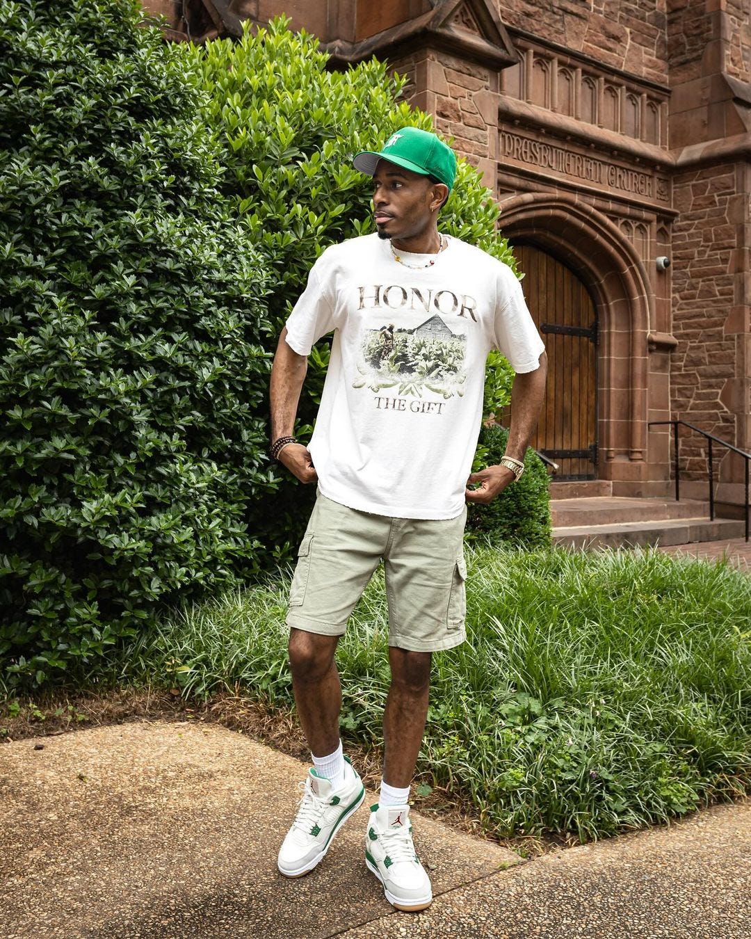 man in front of a church wearing a t-shirt that says "Honor the gift" , cargo shorts, and white Nike sneakers with green accents