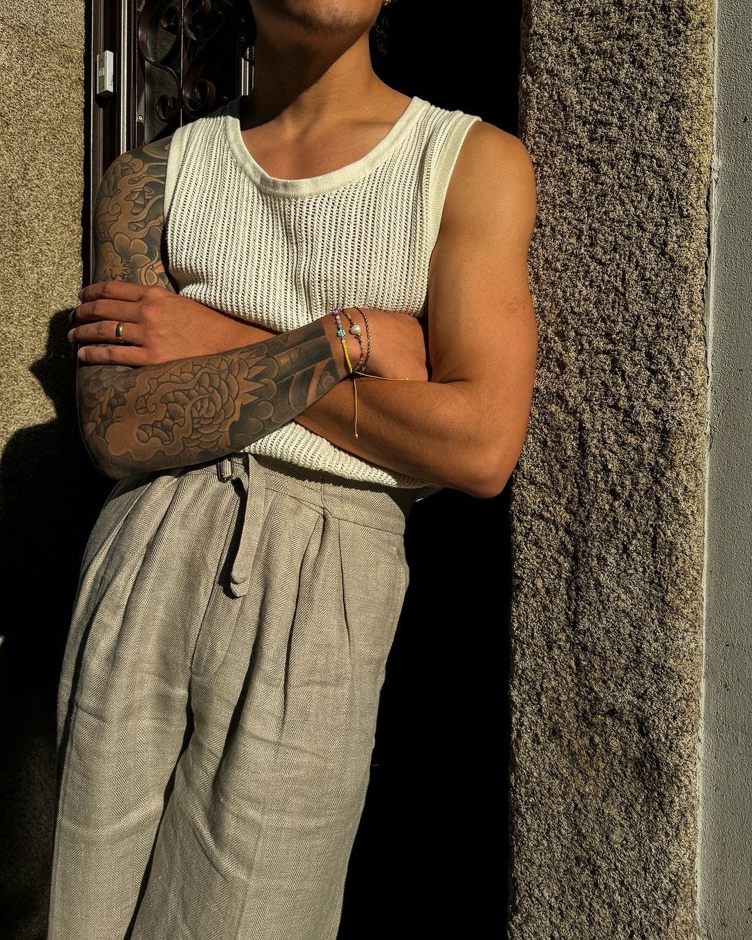 closeup of a man standing in a doorway outside, wearing a white bucket cap, knit sleeveless shirt, and tan linen pants