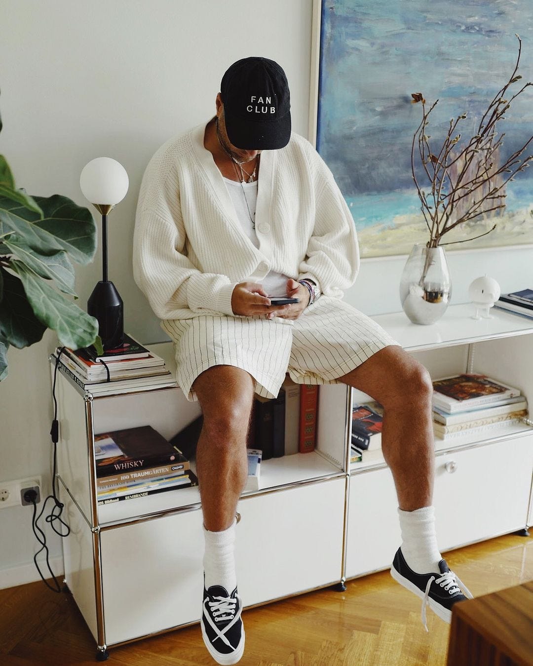 man sitting on a console table wearing an oversized white cardigan with white, striped shorts. he is looking down at his phone