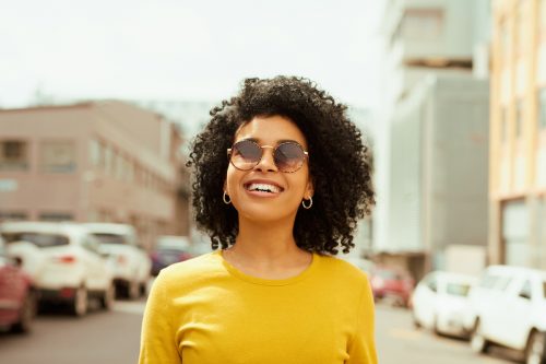 Shot of a young woman looking happy while out in the city; she's wearing a yellow shirt and sunglasses