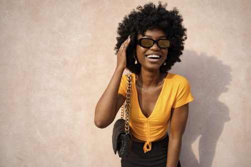 Happy woman wearing a yellow shirt, black pants, and black sunglasses poses against a beige wall