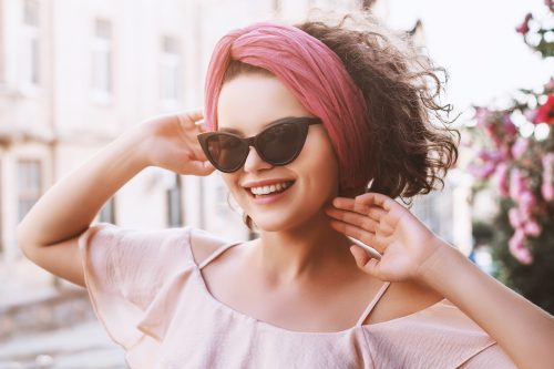 Outdoor close up portrait of happy, young, fashionable woman wearing trendy wide pink headband, black cat eye sunglasses, posing in street of European city