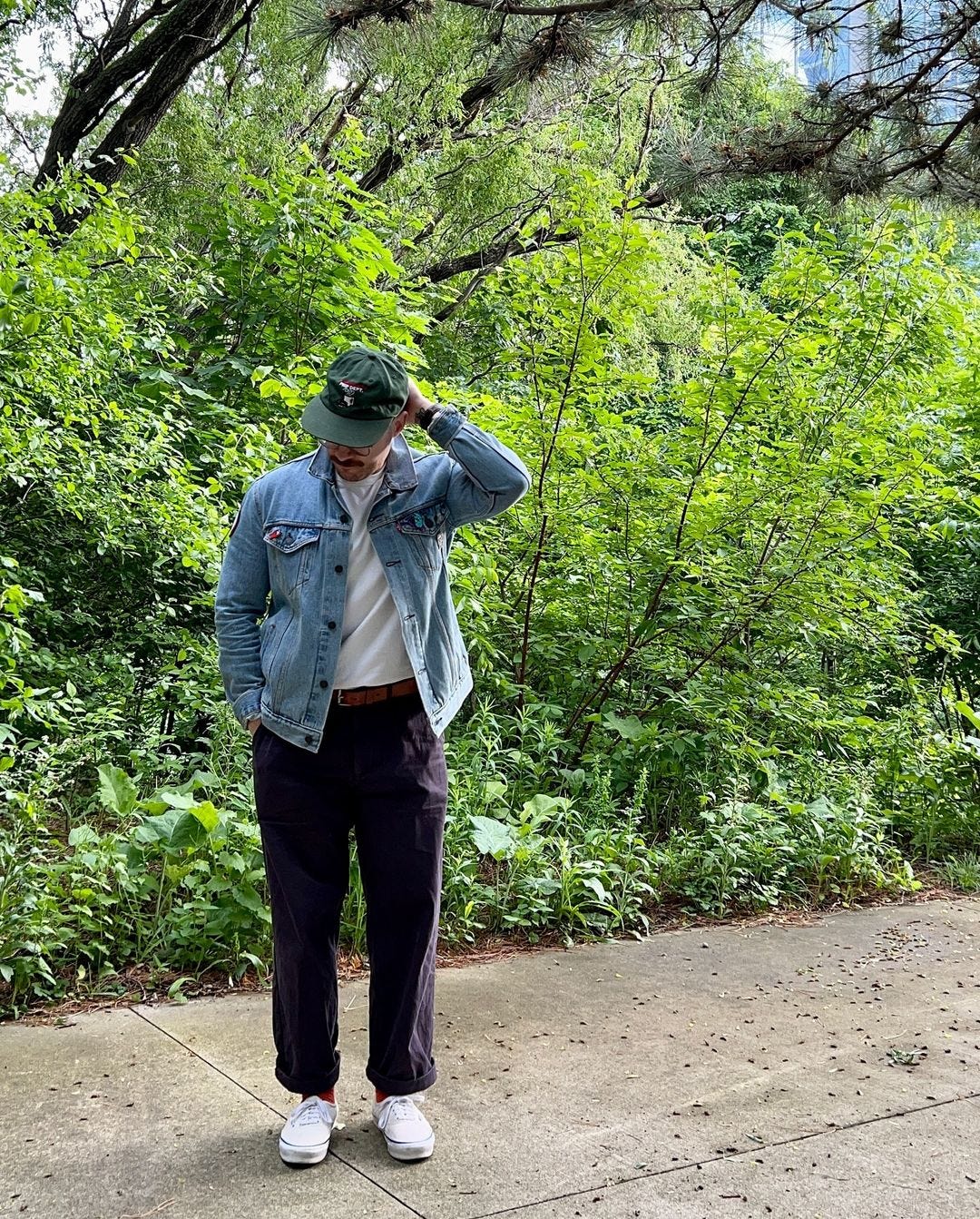 man standing outside in front of greenery wearing a baseball cap, denim jacket, white t-shirt, dark pants, and canvas sneakers