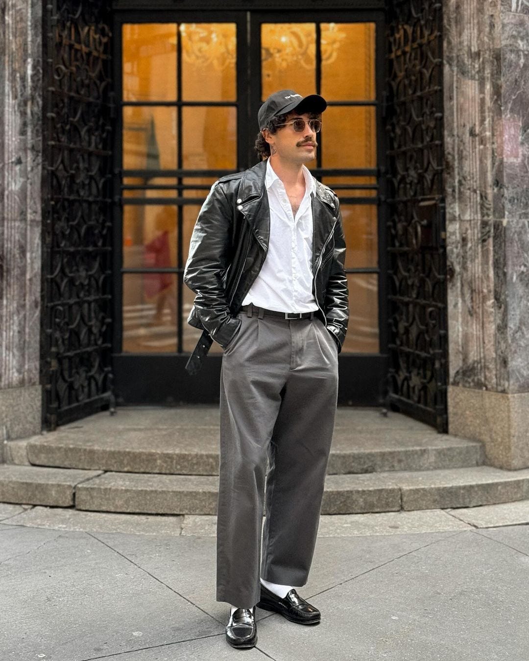 man standing in front of a building wearing a black leather jacket, white button-down shirt, grey slacks, and black loafers with white socks