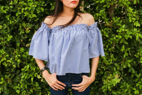 woman wearing a blue-and-white striped off-the-shoulder top and jeans standing against a green plant wall