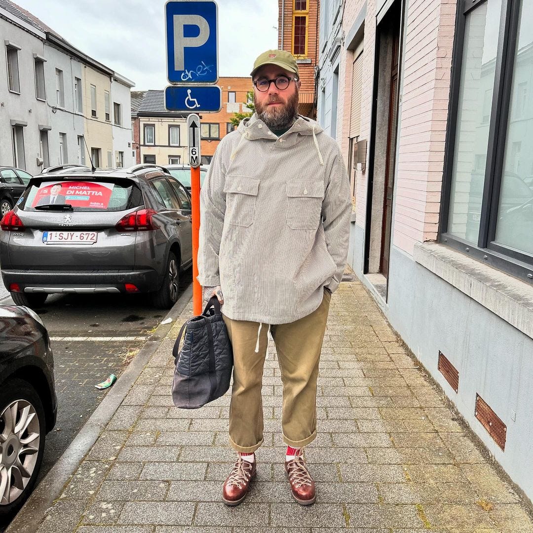 man standing on a city street wearing a striped anorak and khaki pants, holding a work bag