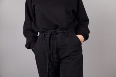 Cropped image of a woman wearing a black sweater and black jeans against a gray background