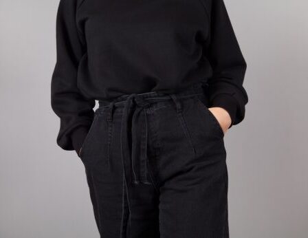 Cropped image of a woman wearing a black sweater and black jeans against a gray background