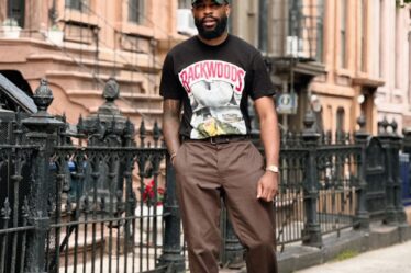 man standing on the sidewalk in front of a brownstone wearing brown pants, black sneakers, and a black t-shirt with a graphic print on the front. he's also wearing a green baseball cap and a watch