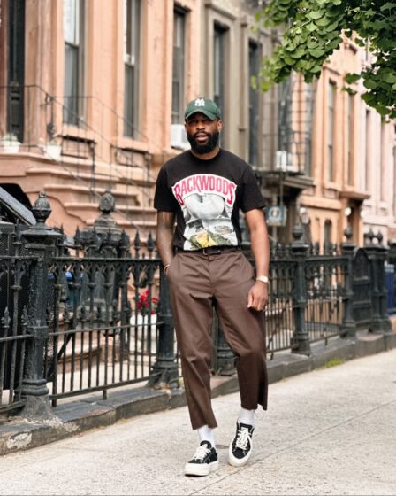 man standing on the sidewalk in front of a brownstone wearing brown pants, black sneakers, and a black t-shirt with a graphic print on the front. he's also wearing a green baseball cap and a watch