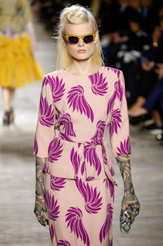 A blonde model in sunglasses wearing a pale pink dress with bright pink angel wings on it.