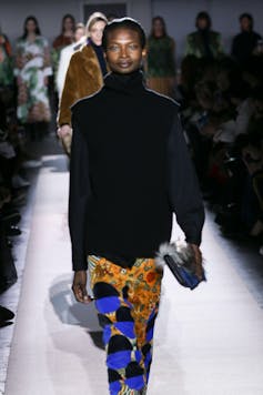 A black model on a catwalk wearing a black jumper and a gold, blue and black skirts.