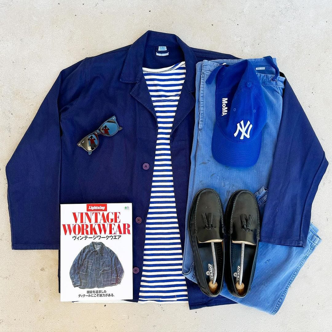 flat lay of a men's outfit featuring a blue chore coat, blue and white striped shirt, blue baseball cap, blue jeans, black loafers, and sunglasses