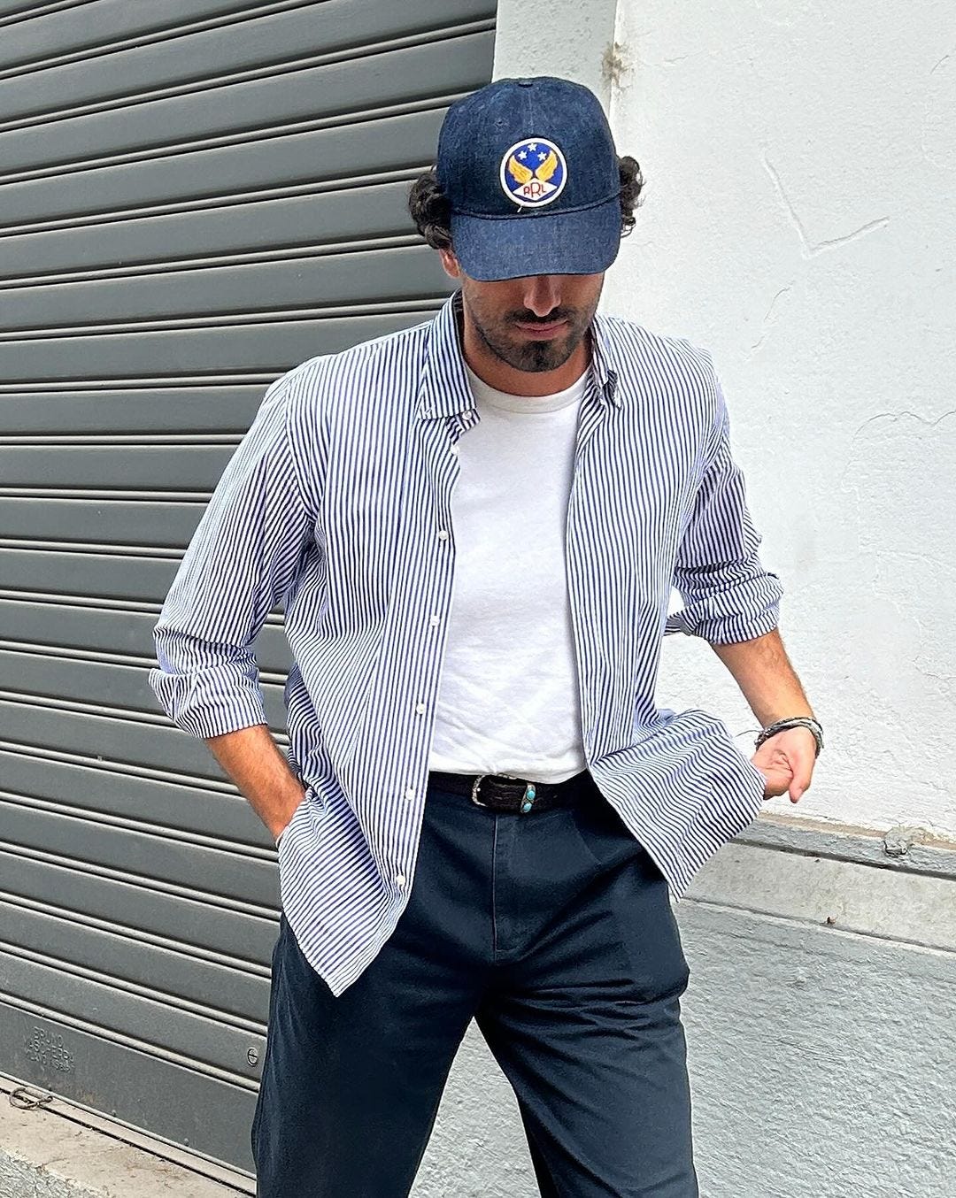 close-up of a man wearing a striped shirt over a white t-shirt with dark blue pants and a blue baseball cap