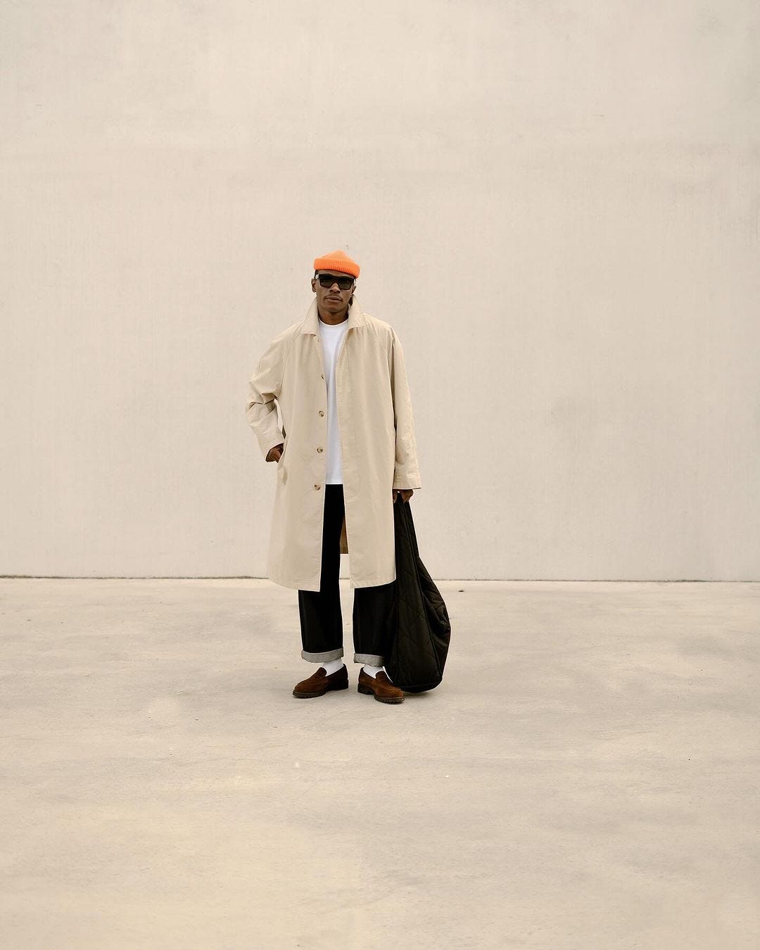 man standing in front of a blank wall, wearing a tan overcoat, white t-shirt, dark jeans, brown loafers, and a small orange beanie