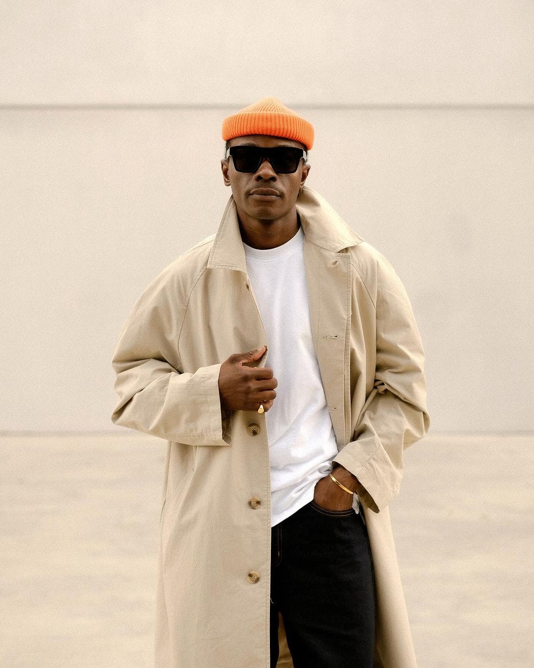 close-up of a man wearing an orange beanie, tan overcoat, white tee, and black pants