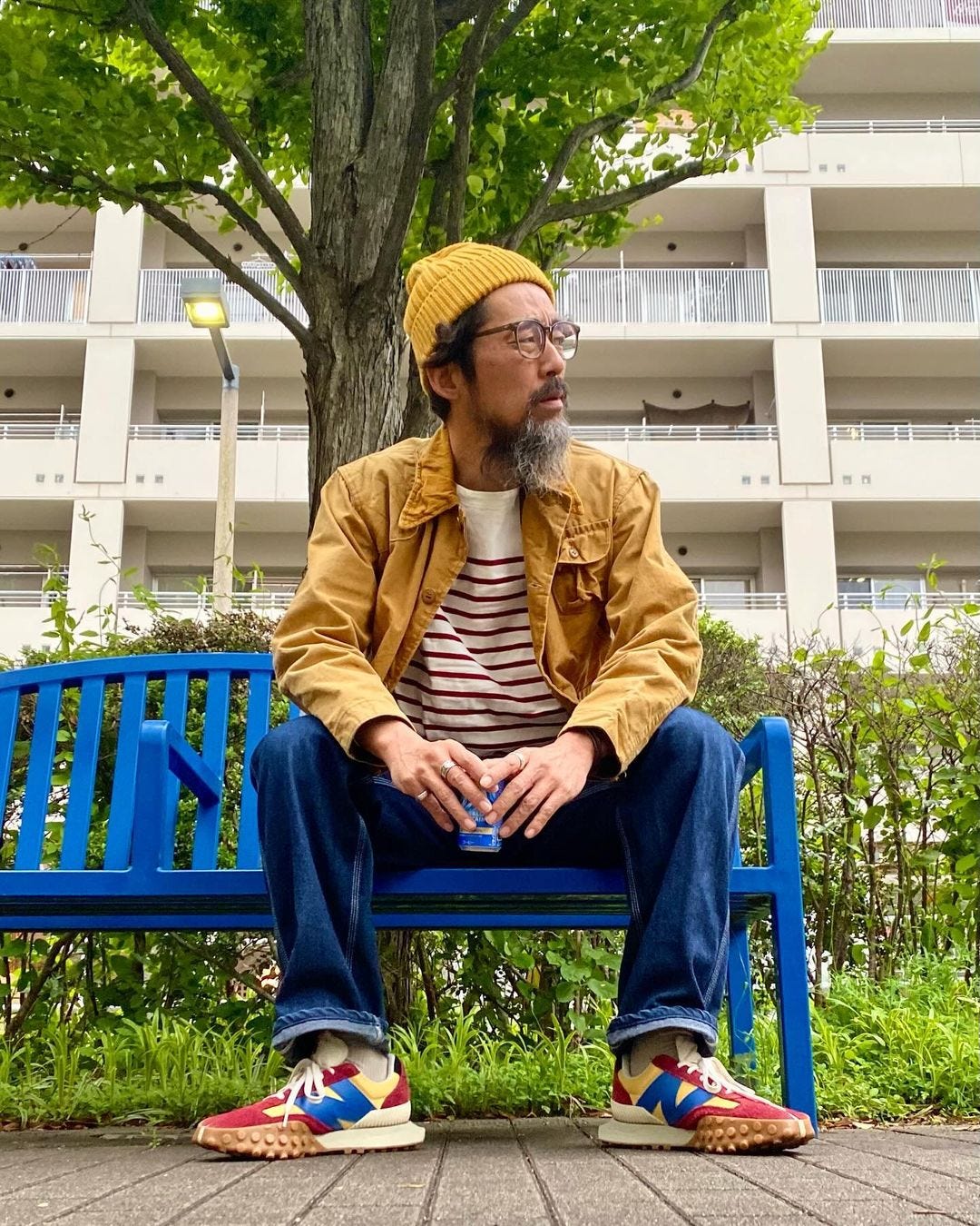 man sitting on a bench in front of a large apartment complex, wearing a yellow beanie, mustard yellow light jacket, red and white striped shirt, blue jeans, and red, blue, and yellow sneakers