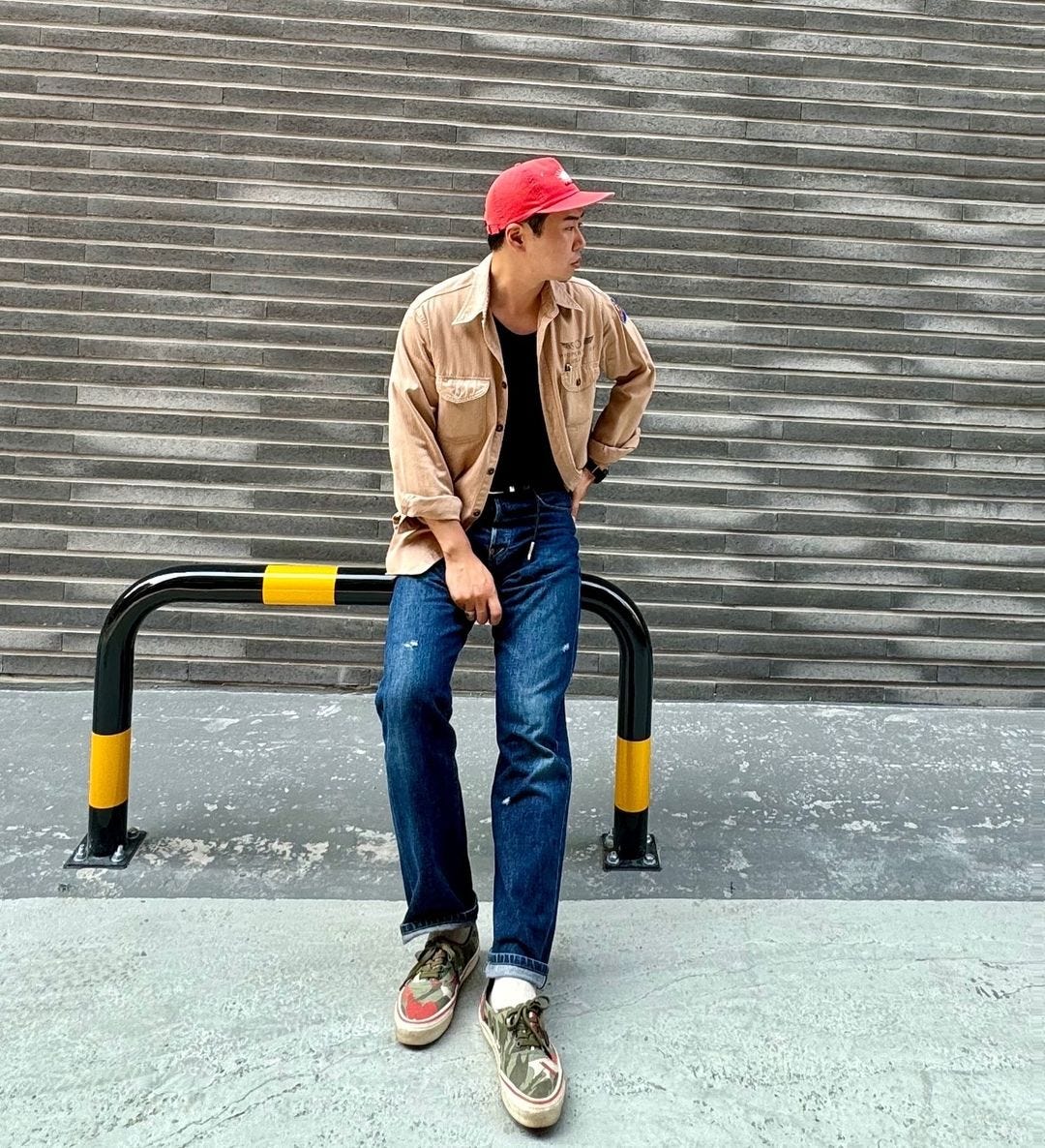 man leaning against a pole, wearing a red baseball cap, tan overshirt, black t-shirt, blue jeans, and green sneakers