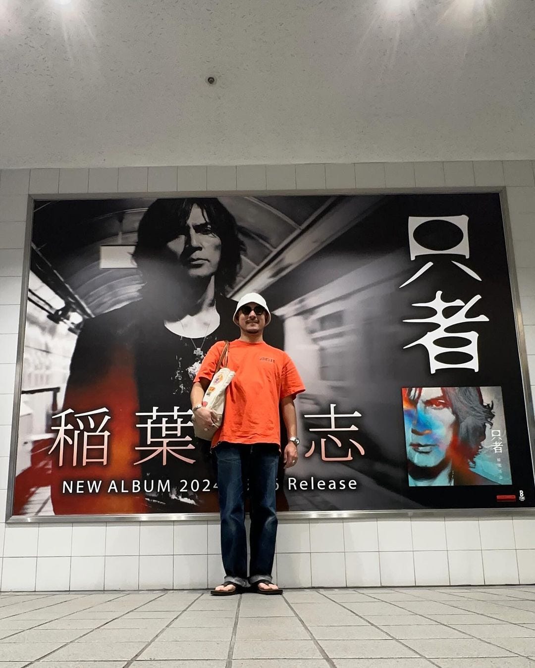 man standing in front of a large advertisement in the subway, wearing an orange t-shirt and dark pants