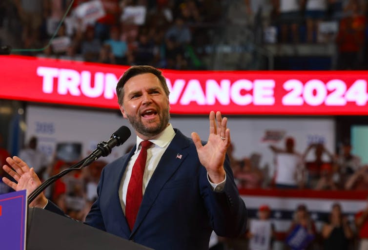 Bearded man spreads arms while speaking at a lectern while wearing a blue suit jacket and scarlet red tie.