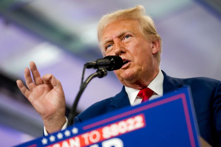 Elderly man with blonde hair and spray tan speaks at microphone while making the 'OK' sign with his right hand.