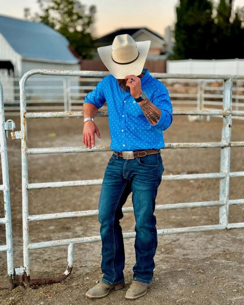 man in a blue shirt and jeans tilting down his cowboy hat