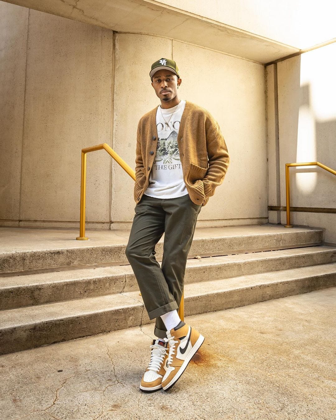 man standing in front of stairs wearing a mustard-colored cardigan, white t-shirt, olive pants, and Nike high top sneakers