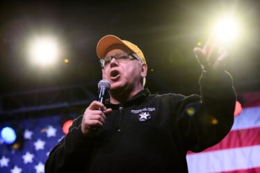 Man wearing glasses and yellow hat holds a microphone and speaks into it while gesturing with his free hand.