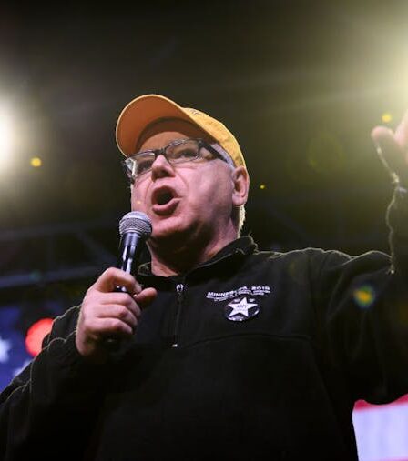 Man wearing glasses and yellow hat holds a microphone and speaks into it while gesturing with his free hand.