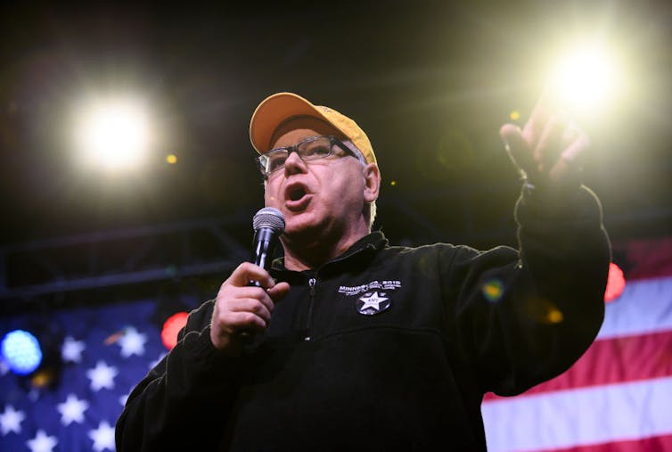 Man wearing glasses and yellow hat holds a microphone and speaks into it while gesturing with his free hand.