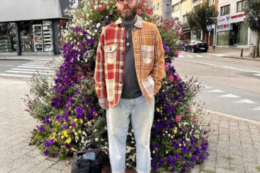 man standing outside in front of a large floral arrangement wearing light jeans and a red plaid overshirt with tan boots