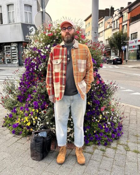 man standing outside in front of a large floral arrangement wearing light jeans and a red plaid overshirt with tan boots