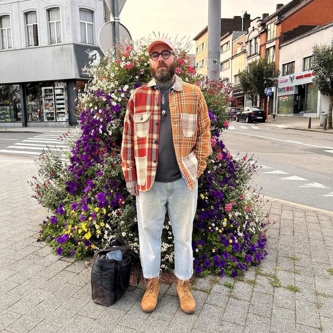 man standing outside in front of a large floral arrangement wearing light jeans and a red plaid overshirt with tan boots