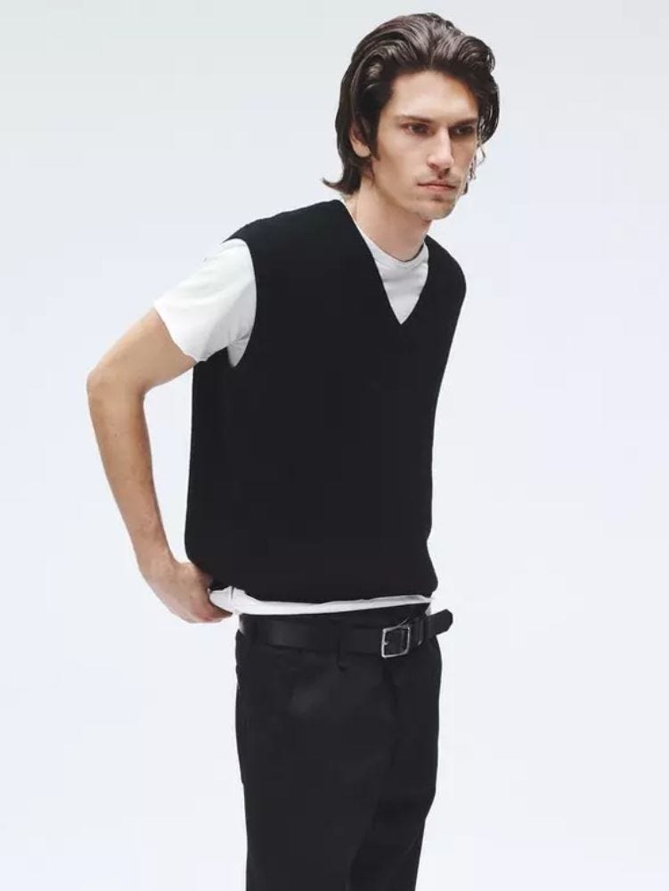 man from the waist up wearing a black v-neck sweater vest over a white t-shirt with black pants and a black belt