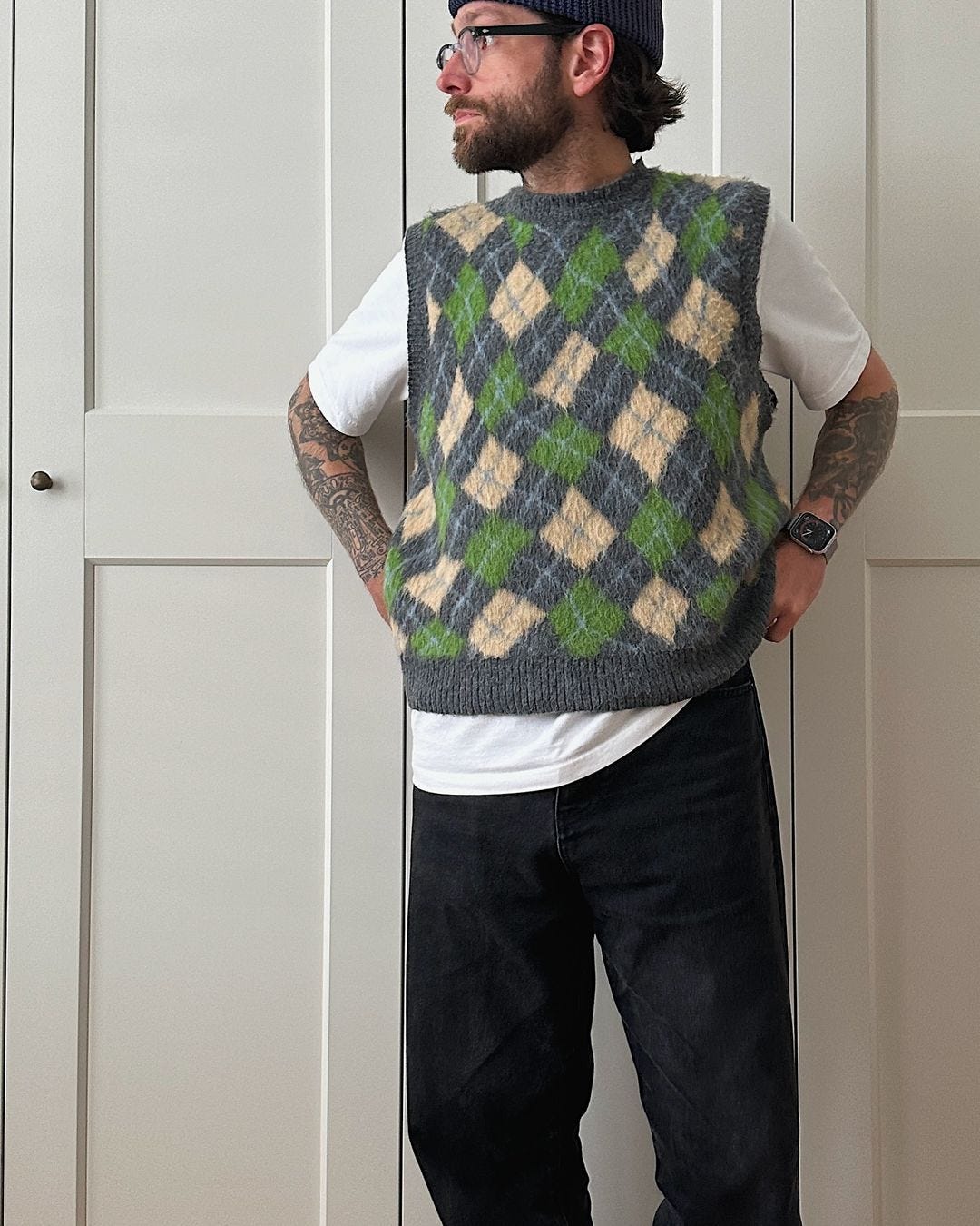 close up of a man wearing glasses, standing in front of closet doors, wearing a patterned green and yellow sweater vest over a white t-shirt with dark pants