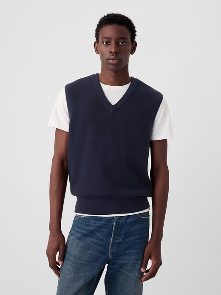 man from the waist up wearing a navy v-neck sweater vest over a white t-shirt with jeans